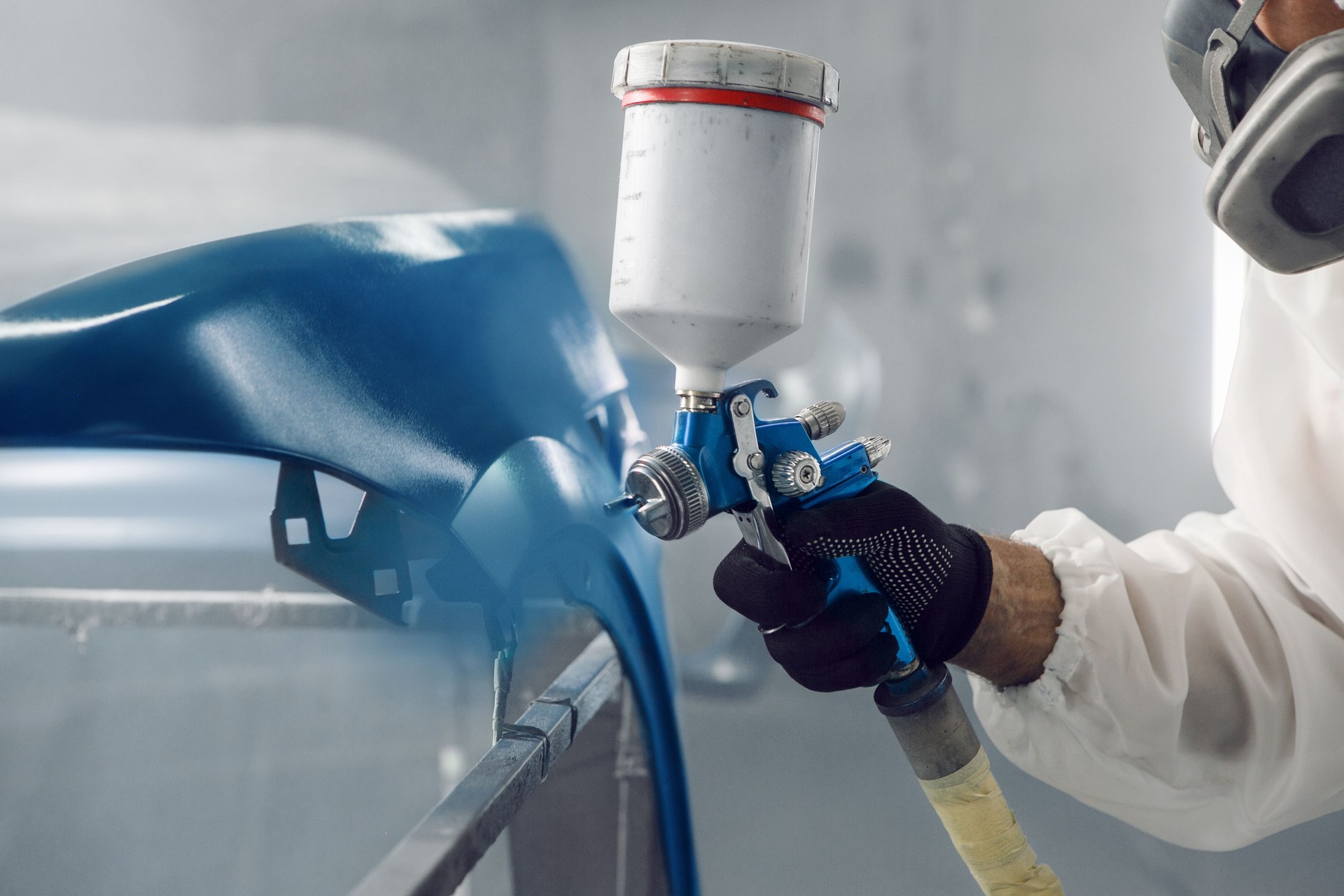 Automobile repairman painter in protective workwear and respirator painting part of car in paint chamber.